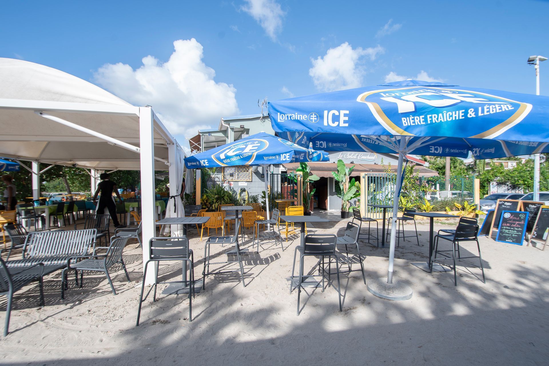 Restaurant en plein air avec tables, chaises et parasols bleus sous un ciel ensoleillé.