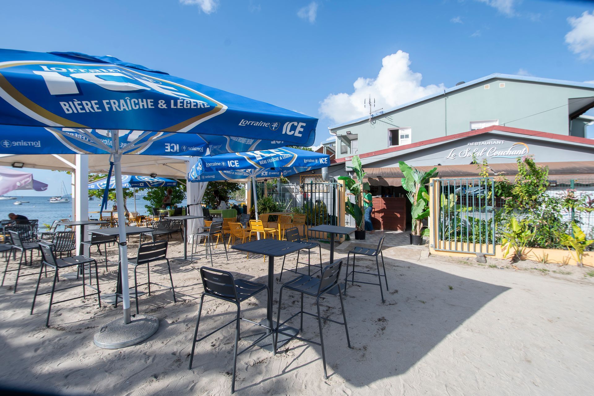 Restaurant de plage en plein air avec tables, chaises et parasols bleus, par une journée ensoleillée.