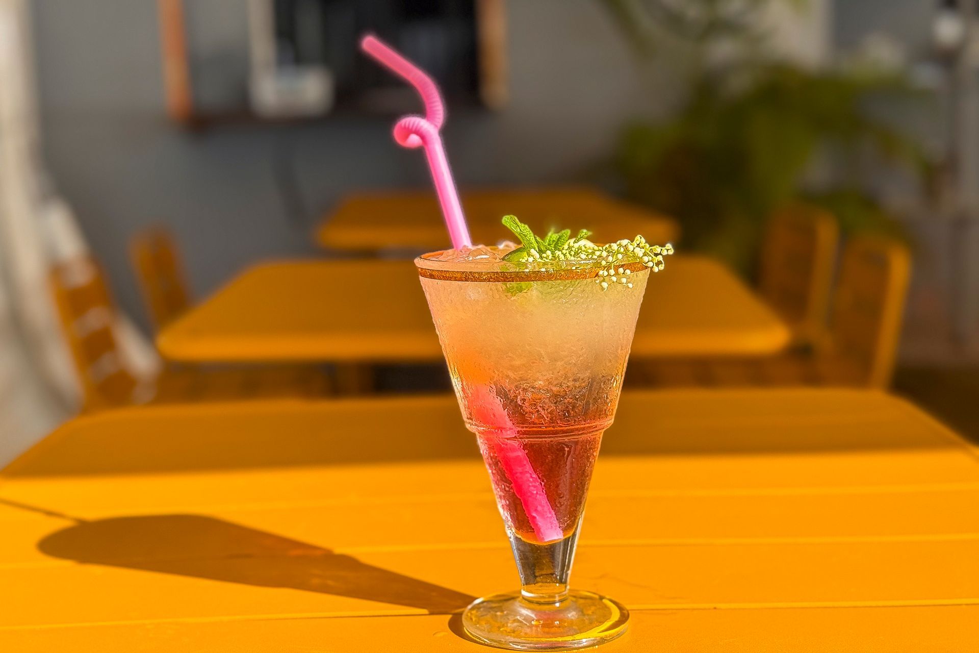 Cocktail servi dans un verre avec une paille rose sur une table jaune, en extérieur.