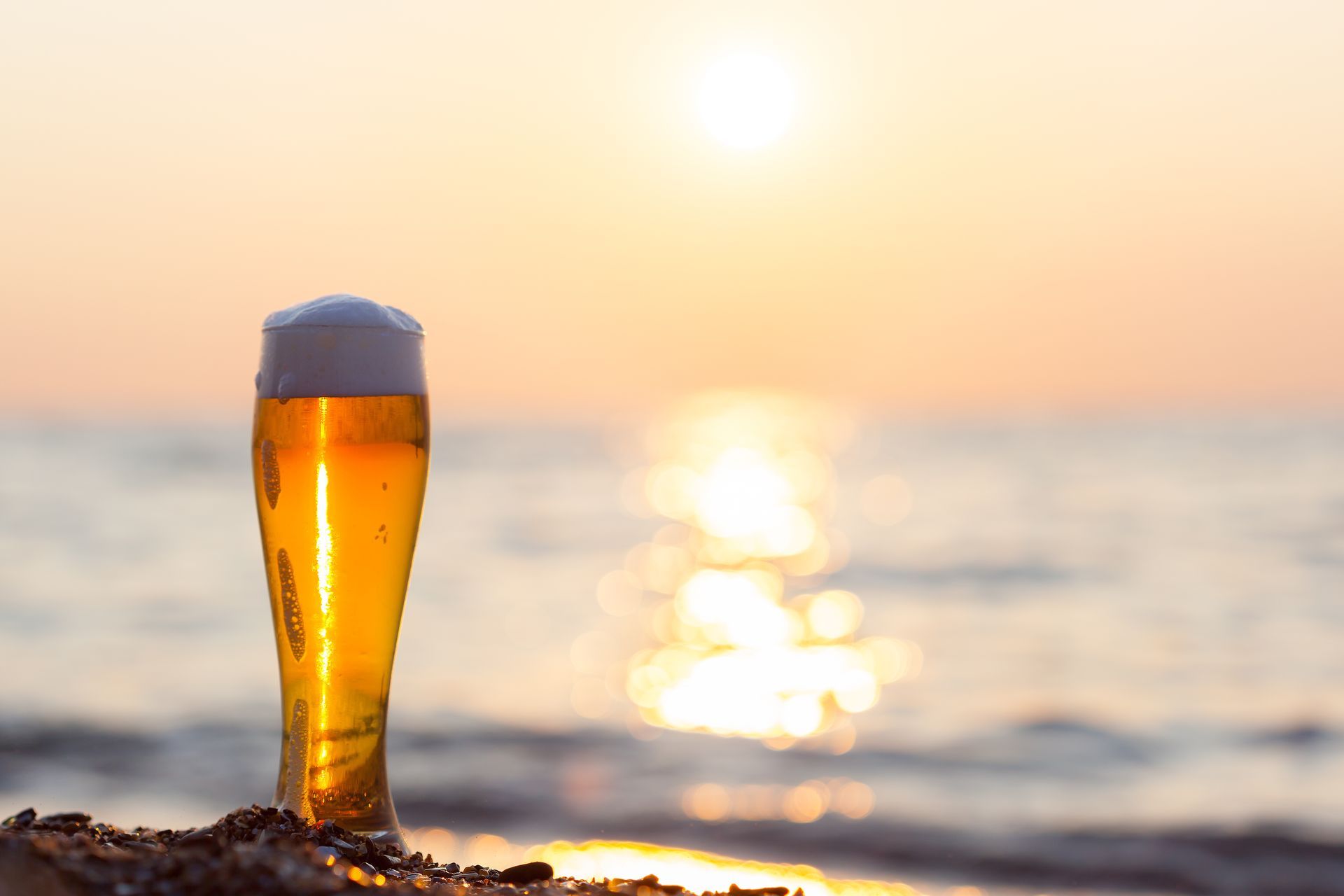Un verre de bière sur une plage de sable au coucher du soleil, avec l'océan en arrière-plan.
