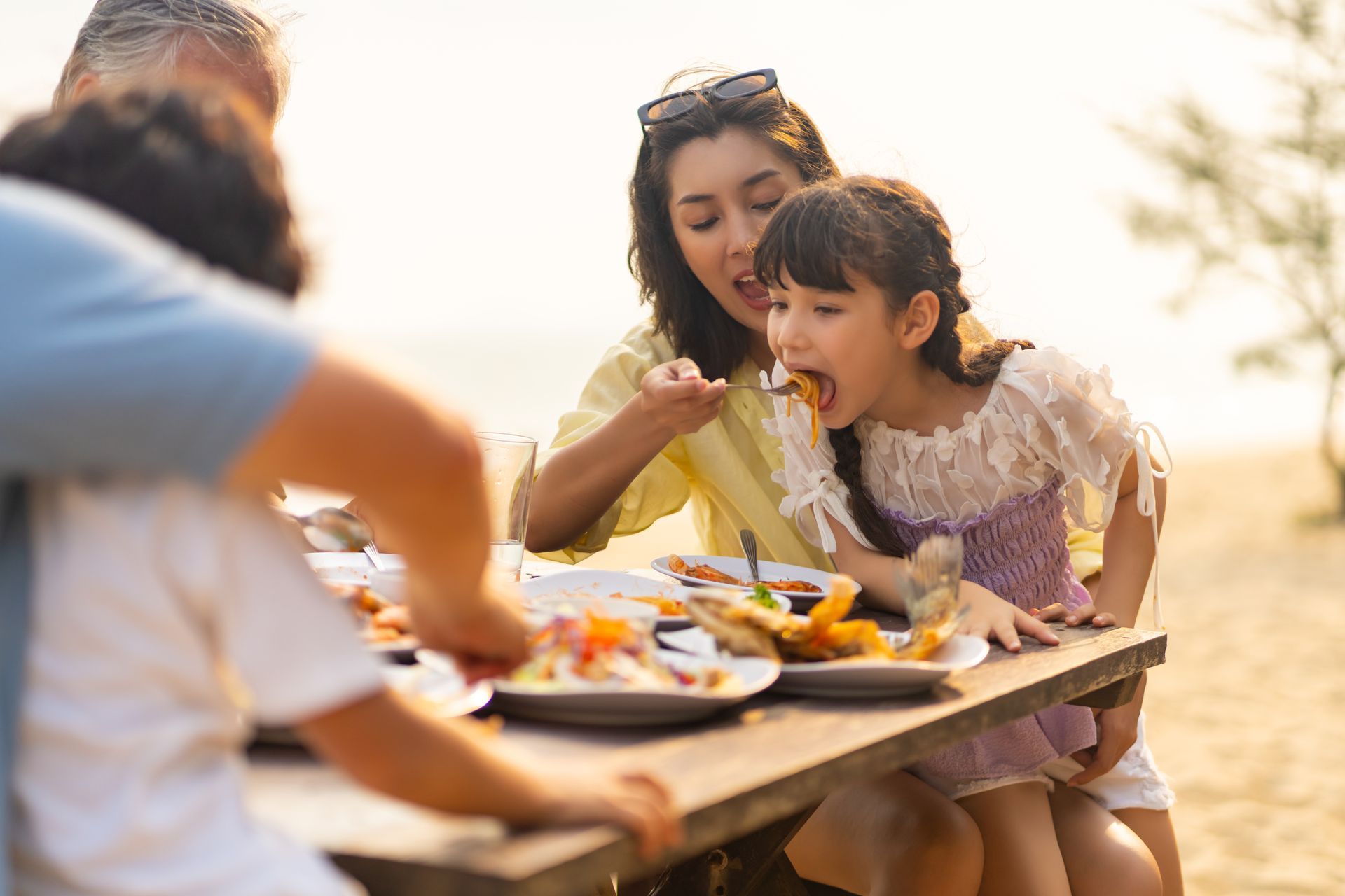 Une famille prend un repas à une table en bord de mer ; une femme donne à manger à une fillette, d'autres se servent.