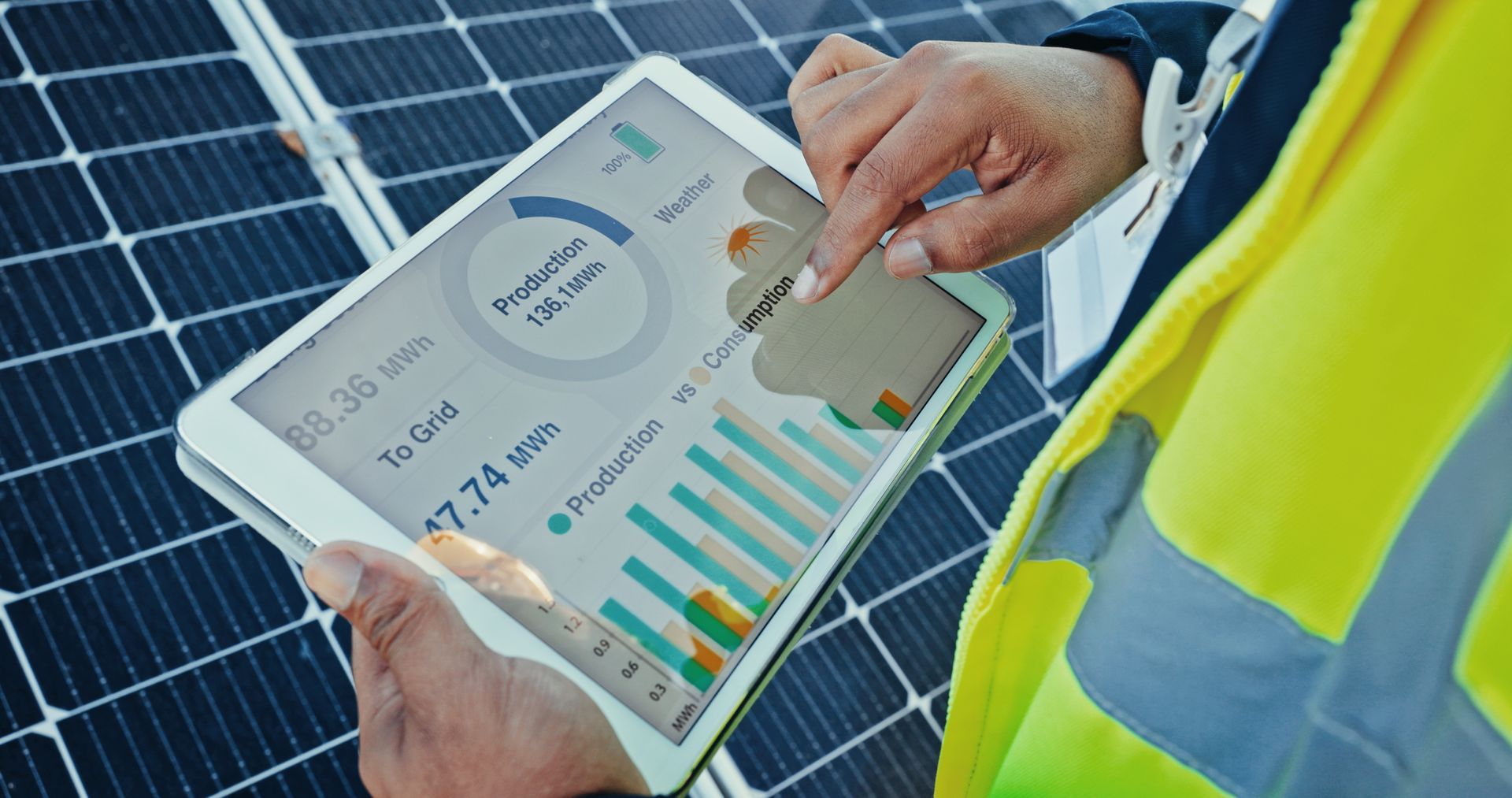 Une personne portant un gilet à haute visibilité tient une tablette affichant des données de production d'énergie solaire devant un ensemble de panneaux solaires.