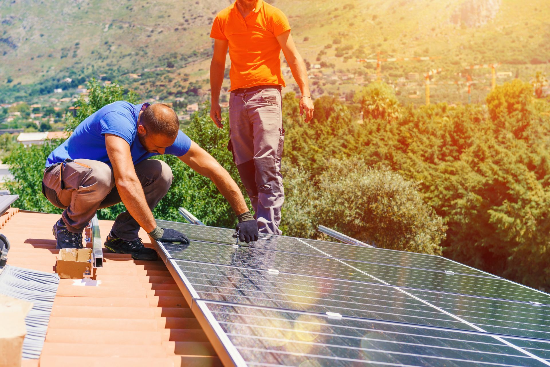 Deux ouvriers en tenue de travail installent des panneaux solaires sur un toit de tuiles en terre cuite sous un soleil éclatant.