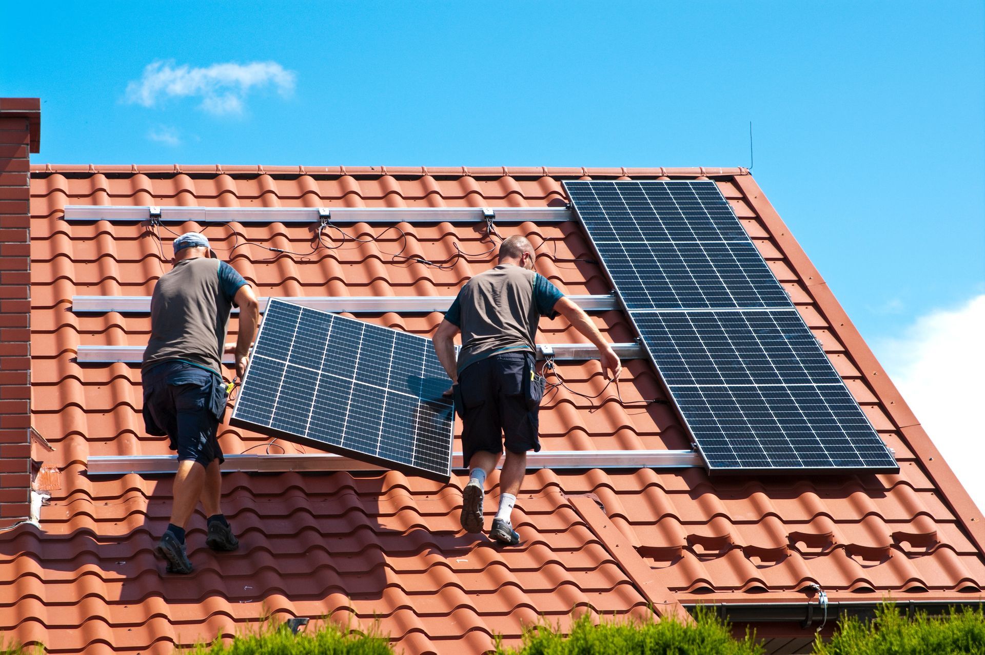 Deux personnes installent des panneaux solaires sur un toit de tuiles rouges, sur fond de ciel bleu azur.