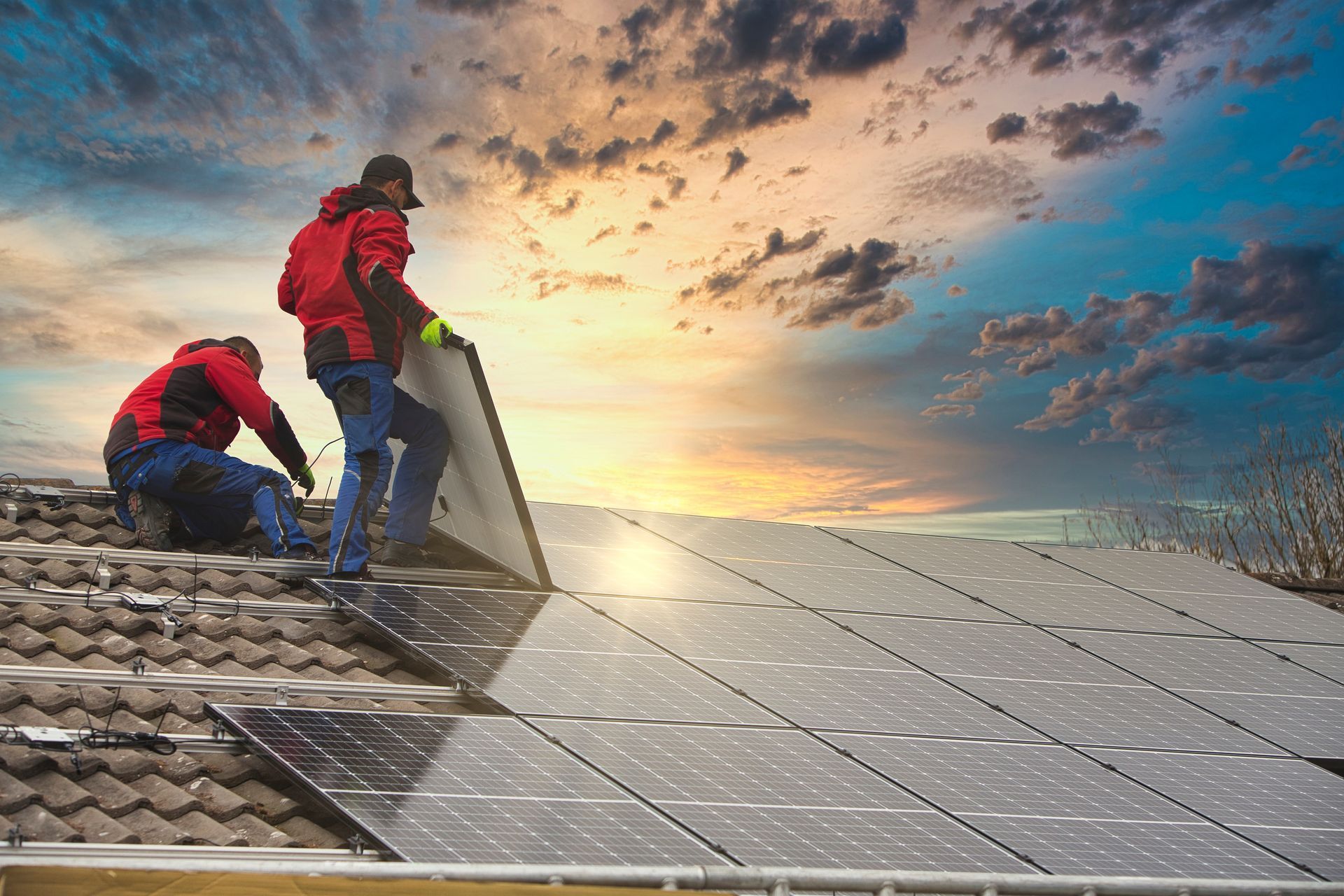 Deux ouvriers en vestes rouges installent des panneaux solaires sur un toit de tuiles au coucher du soleil.