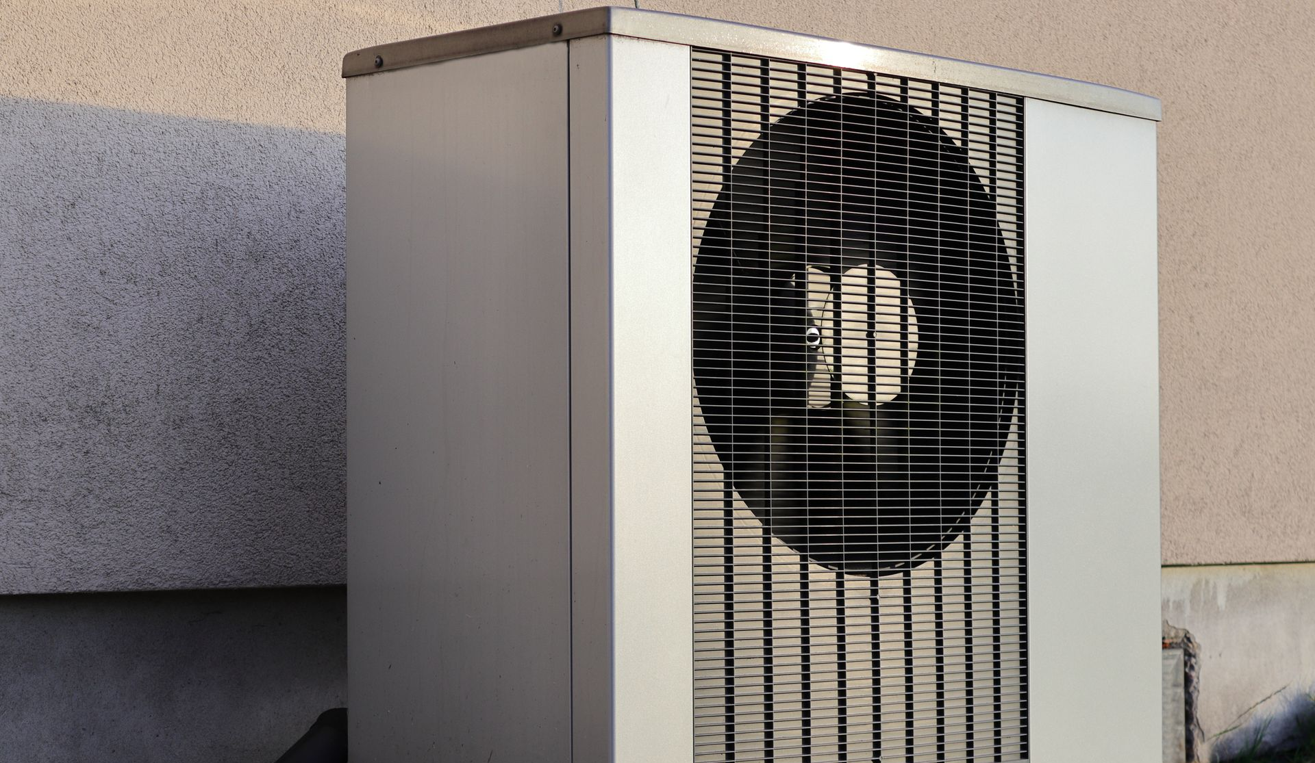 Unité de pompe à chaleur extérieure rectangulaire et métallique, dotée d'une grande grille de ventilation circulaire, placée contre un mur de couleur neutre.