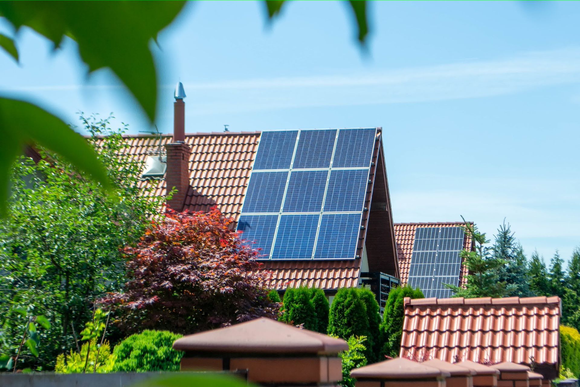 Un toit d'habitation recouvert de panneaux solaires, avec des arbres verts et une clôture en briques rouges au premier plan, sous un ciel bleu.