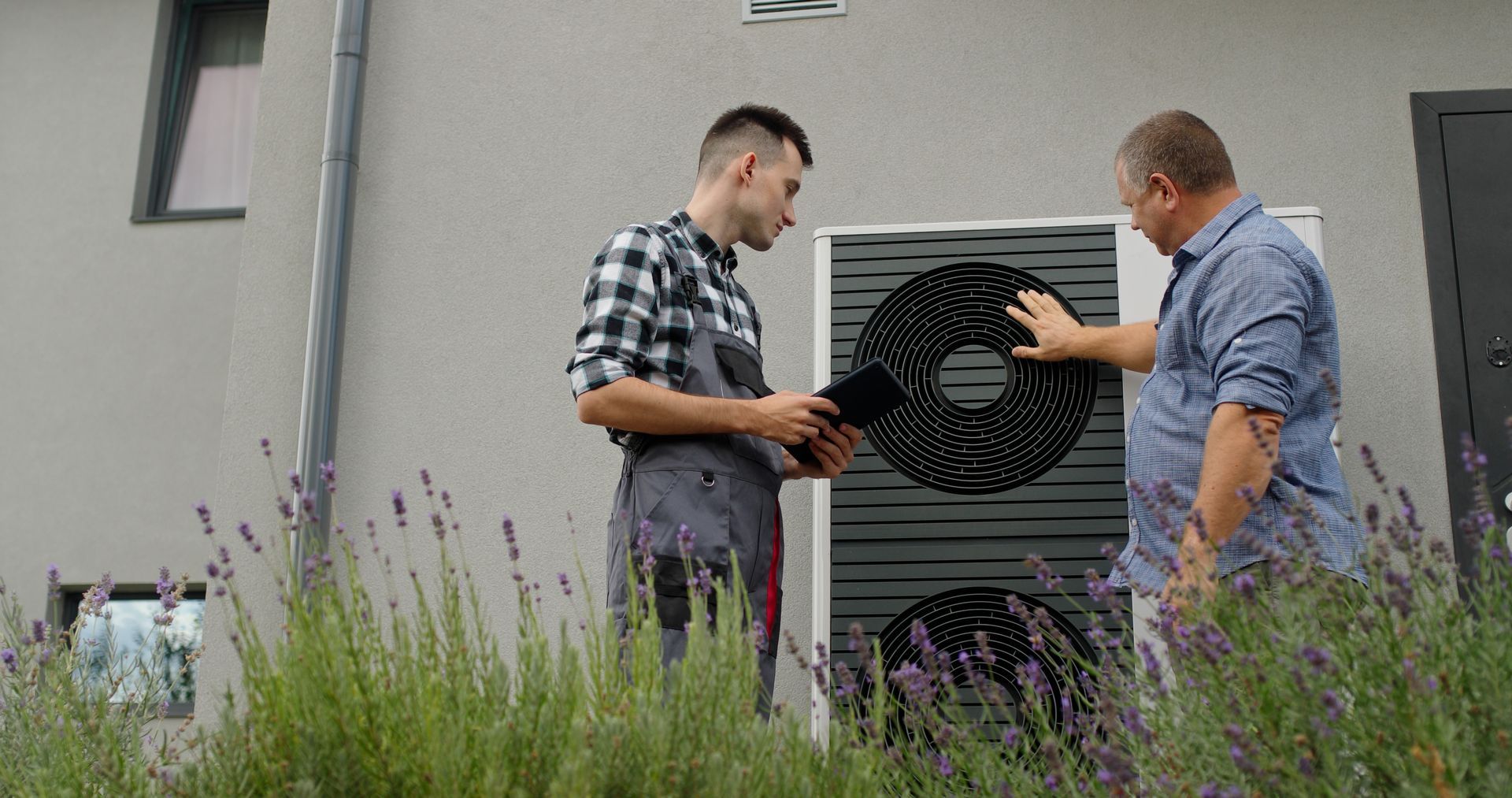 Un technicien inspecte une pompe à chaleur à l'extérieur avec un client, près de buissons de lavande adossés à un mur gris.