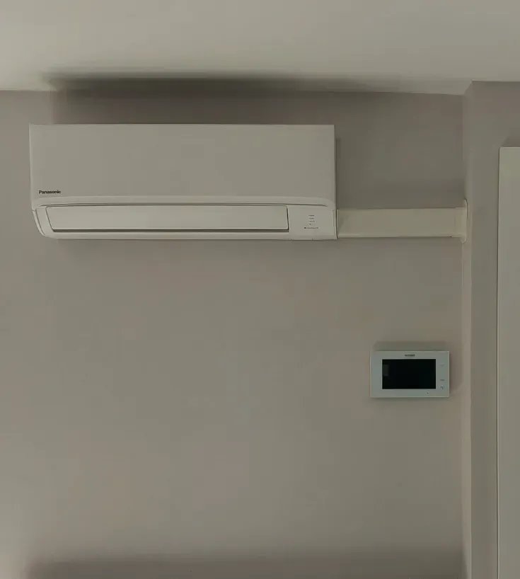 Un climatiseur mural blanc à côté d'une petite commande murale numérique rectangulaire.