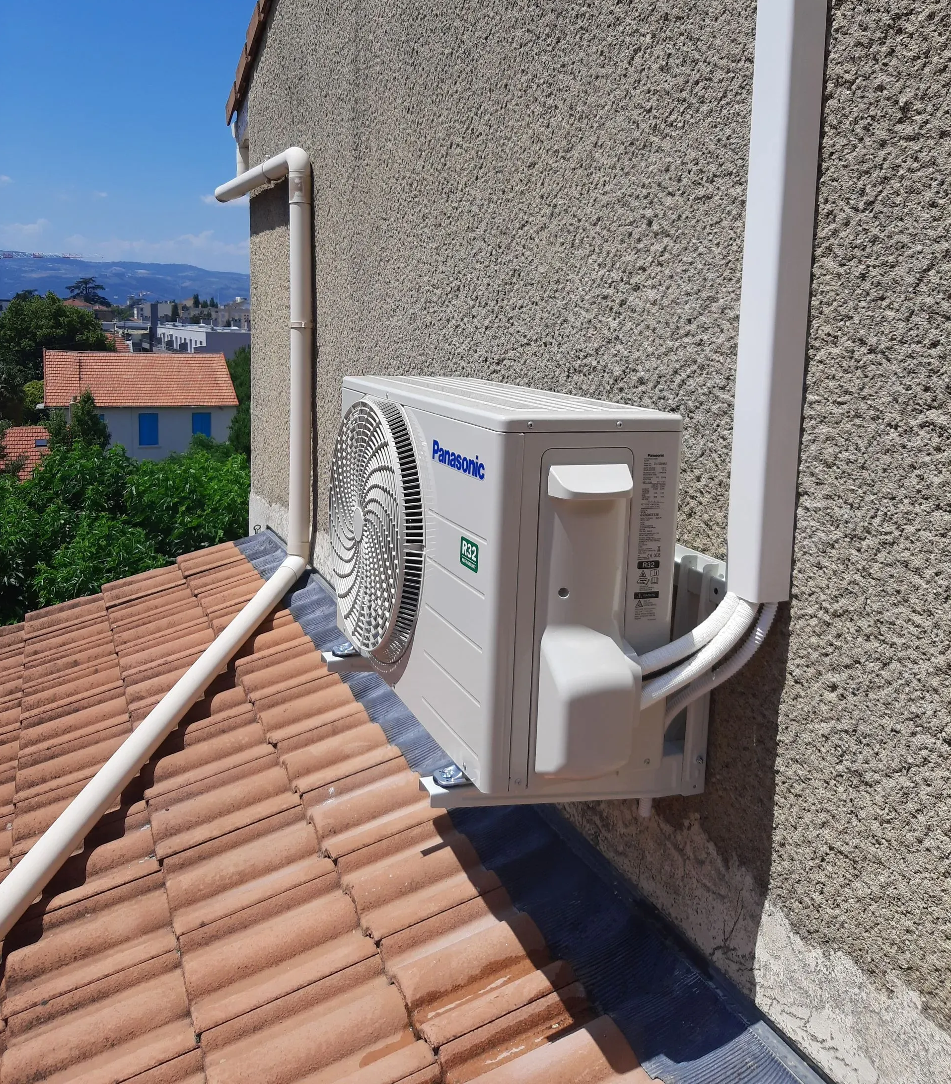 Un climatiseur extérieur fixé sur un mur beige au-dessus d'un toit de tuiles, sous un ciel bleu dégagé.