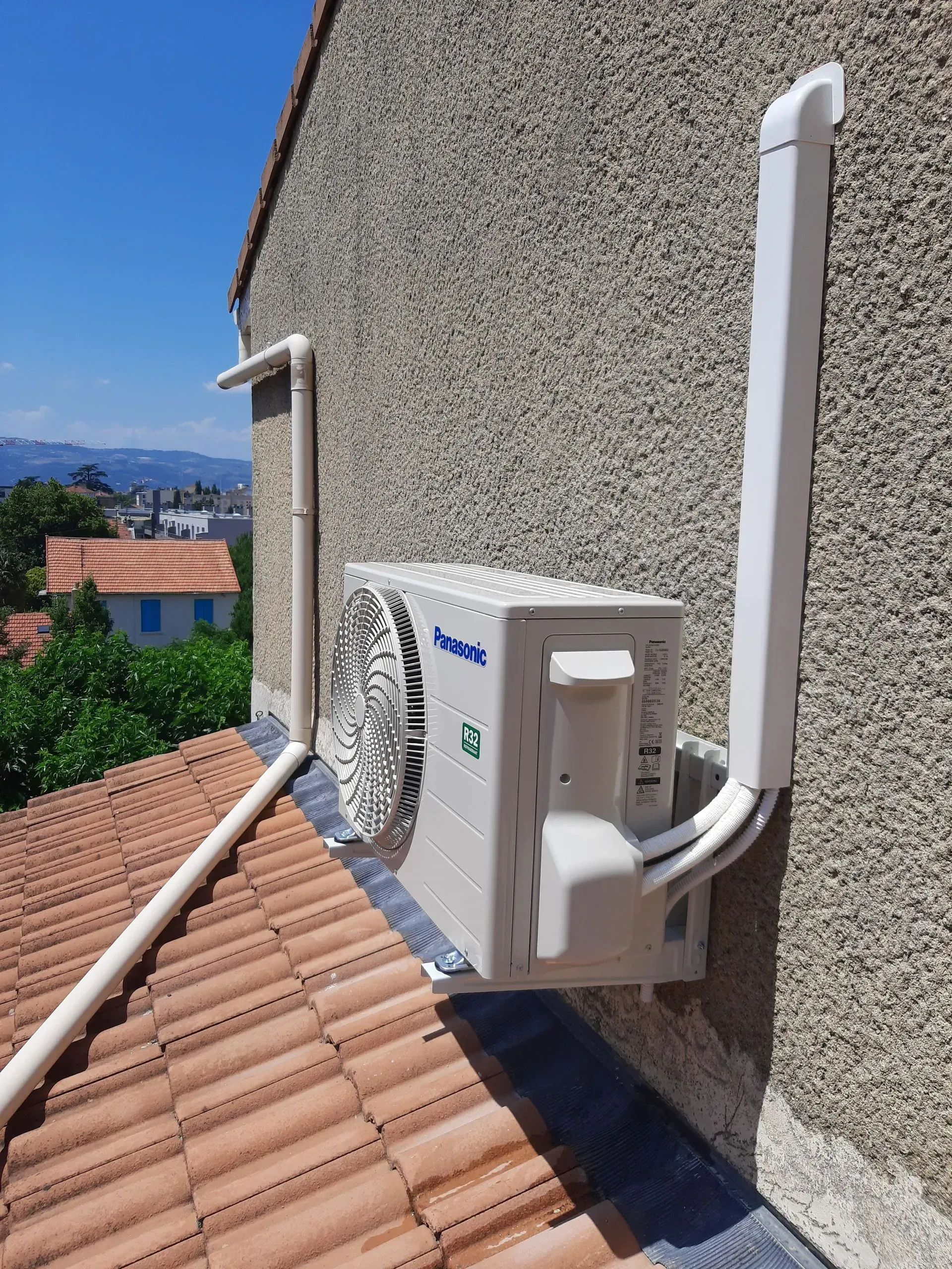 Unité de climatisation extérieure blanche, montée sur un mur texturé au-dessus d'un toit de tuiles, avec des conduits de protection blancs installés.