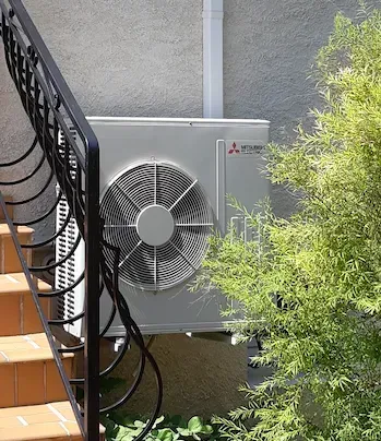 Une unité de climatisation extérieure Mitsubishi est installée sur un piédestal à côté d'un mur en stuc beige et d'un escalier en métal noir.