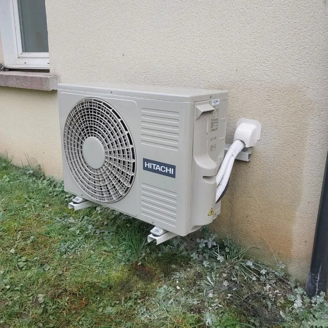 Un climatiseur extérieur Hitachi beige, monté sur un mur extérieur clair au-dessus d'une parcelle de pelouse verte.