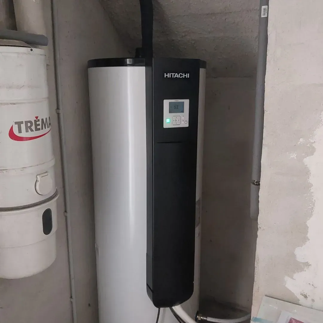 Un chauffe-eau thermodynamique Hitachi avec un panneau de commande noir se trouve dans un sous-sol à côté d'un réservoir Trema blanc.