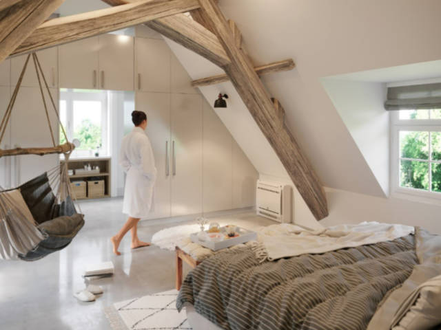 Une personne en peignoir traverse une chambre mansardée avec des poutres en bois, un fauteuil suspendu et un lit.