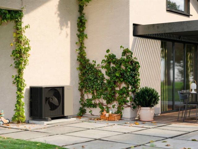 Une pompe à chaleur extérieure est installée sur une terrasse en pierre claire, à côté d'un mur de maison orné de lierre grimpant.