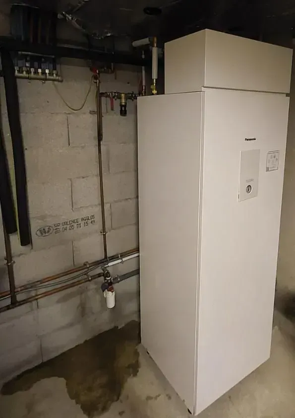 Un chauffe-eau blanc est installé dans un sous-sol, adossé à un mur en parpaings de béton, avec des tuyaux de plomberie en cuivre apparents.