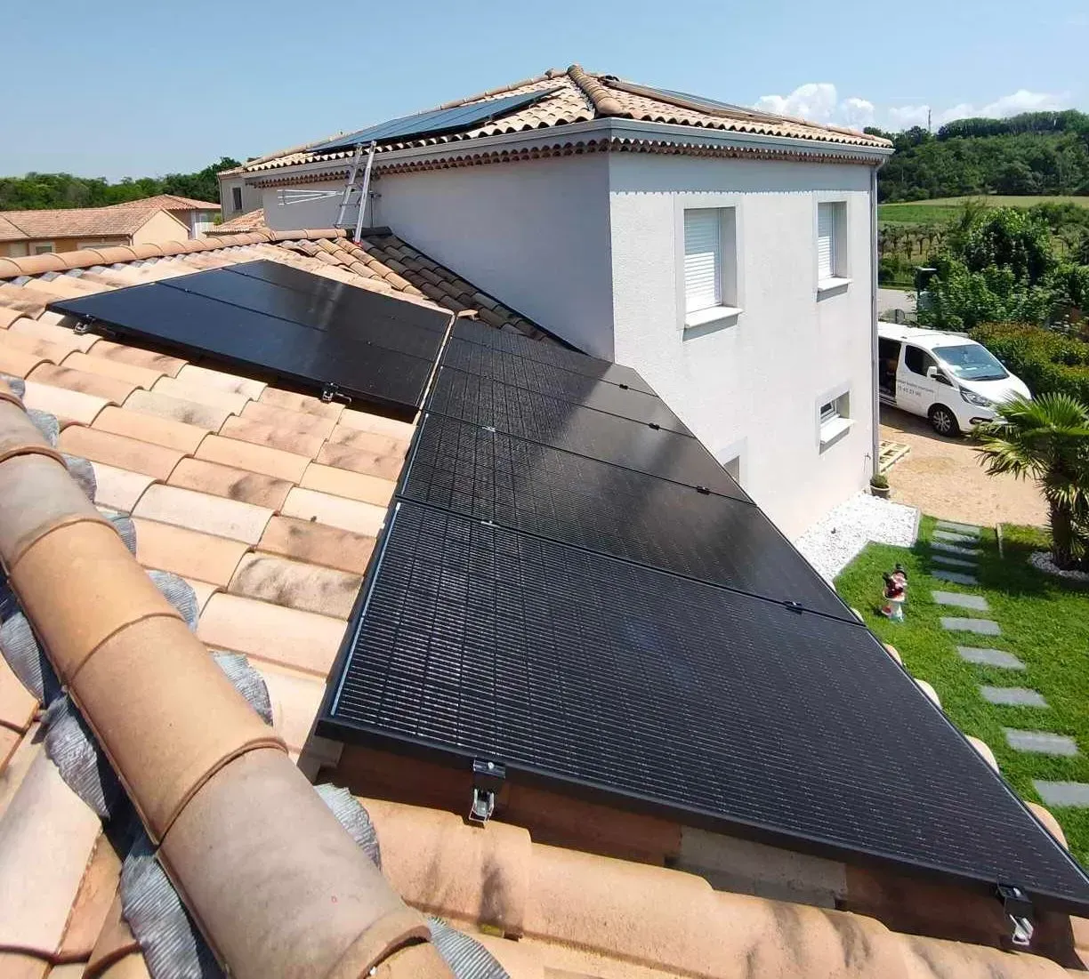 Panneaux solaires noirs installés sur le toit en tuiles de terre cuite d'une maison d'habitation, avec une maison blanche et une camionnette en arrière-plan.