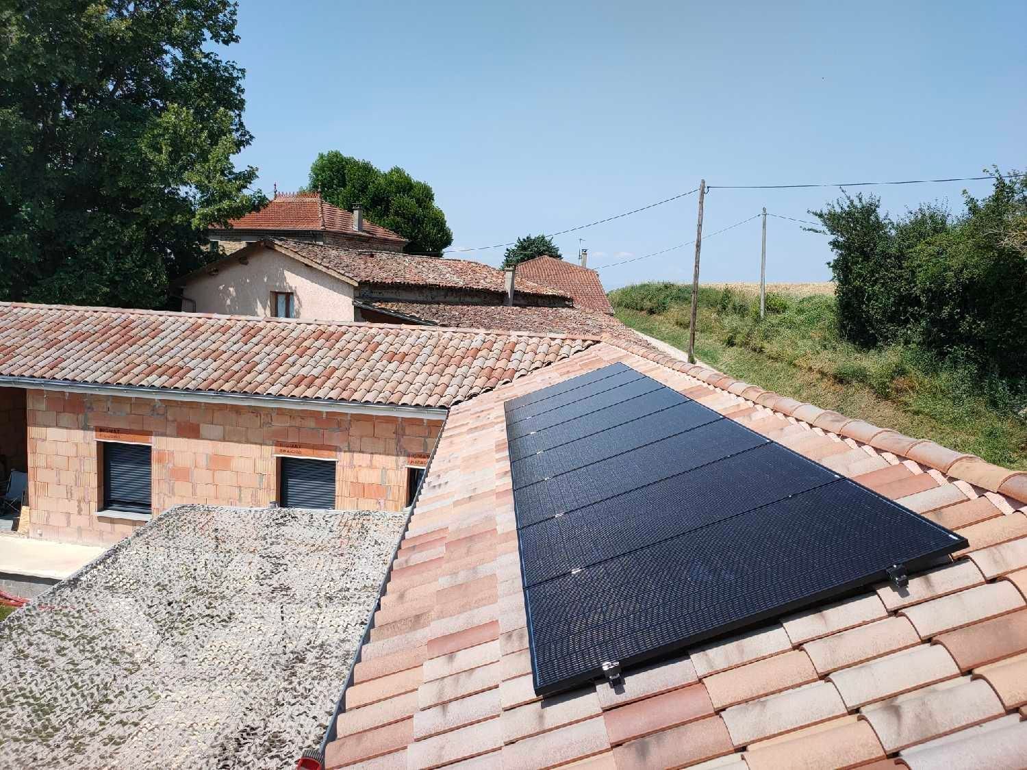 Un panneau solaire rectangulaire installé sur un toit de tuiles en terre cuite, à côté d'un bâtiment rustique en briques, en milieu rural.