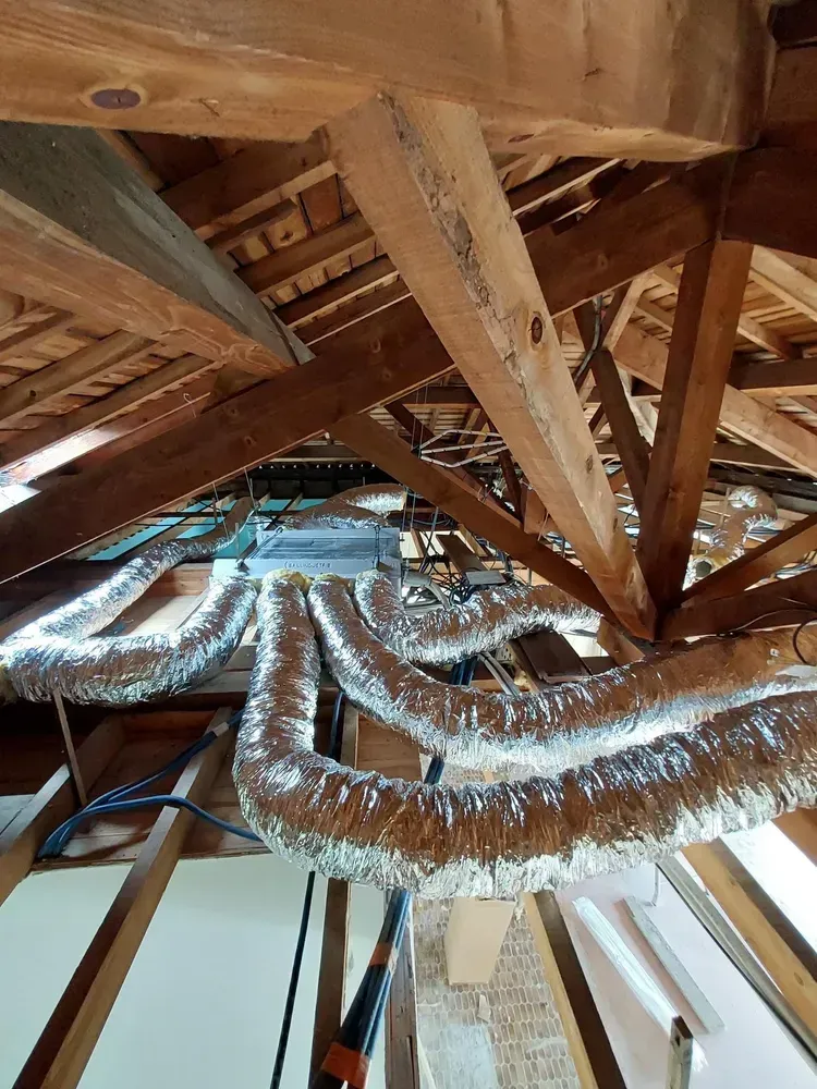 Des conduits de climatisation flexibles argentés traversent une charpente de combles en bois avec des chevrons et des poutres apparents.