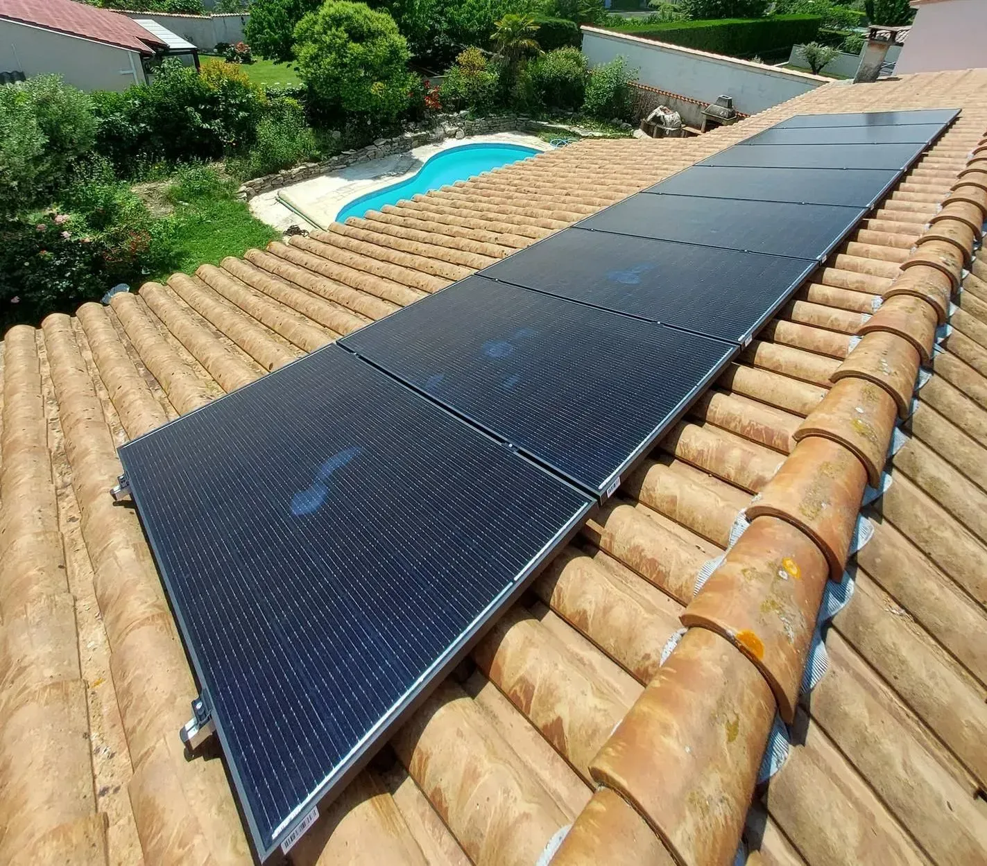 Une rangée de panneaux solaires noirs chauffants pour piscine, installés sur un toit de tuiles en terre cuite donnant sur une piscine dans un jardin.