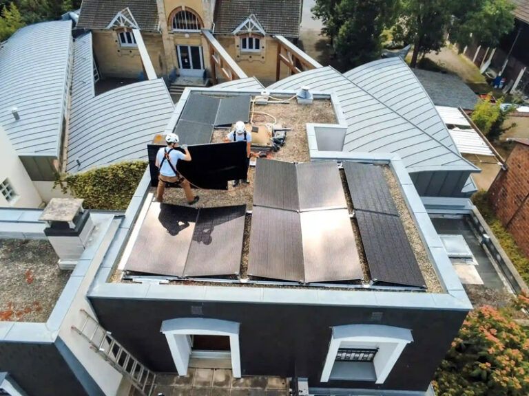 Deux ouvriers, harnachés de sécurité, installent des panneaux solaires sombres sur un toit plat, vus du dessus.