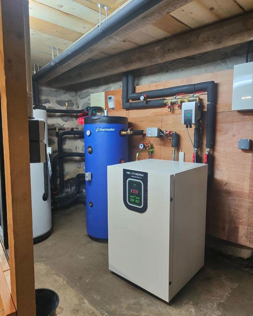 Une salle des machines abritant une pompe à chaleur blanche, un grand réservoir d'eau chaude bleu et une tuyauterie isolée contre un mur en contreplaqué.