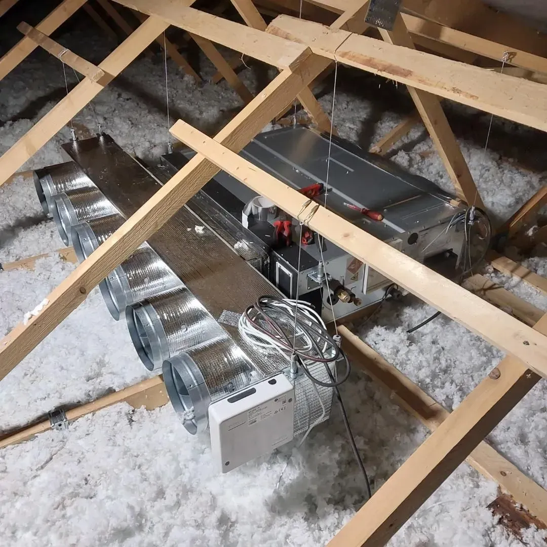 Unité de traitement d'air CVC avec conduits métalliques, installée dans un grenier avec charpente en bois et isolation en fibre de verre.