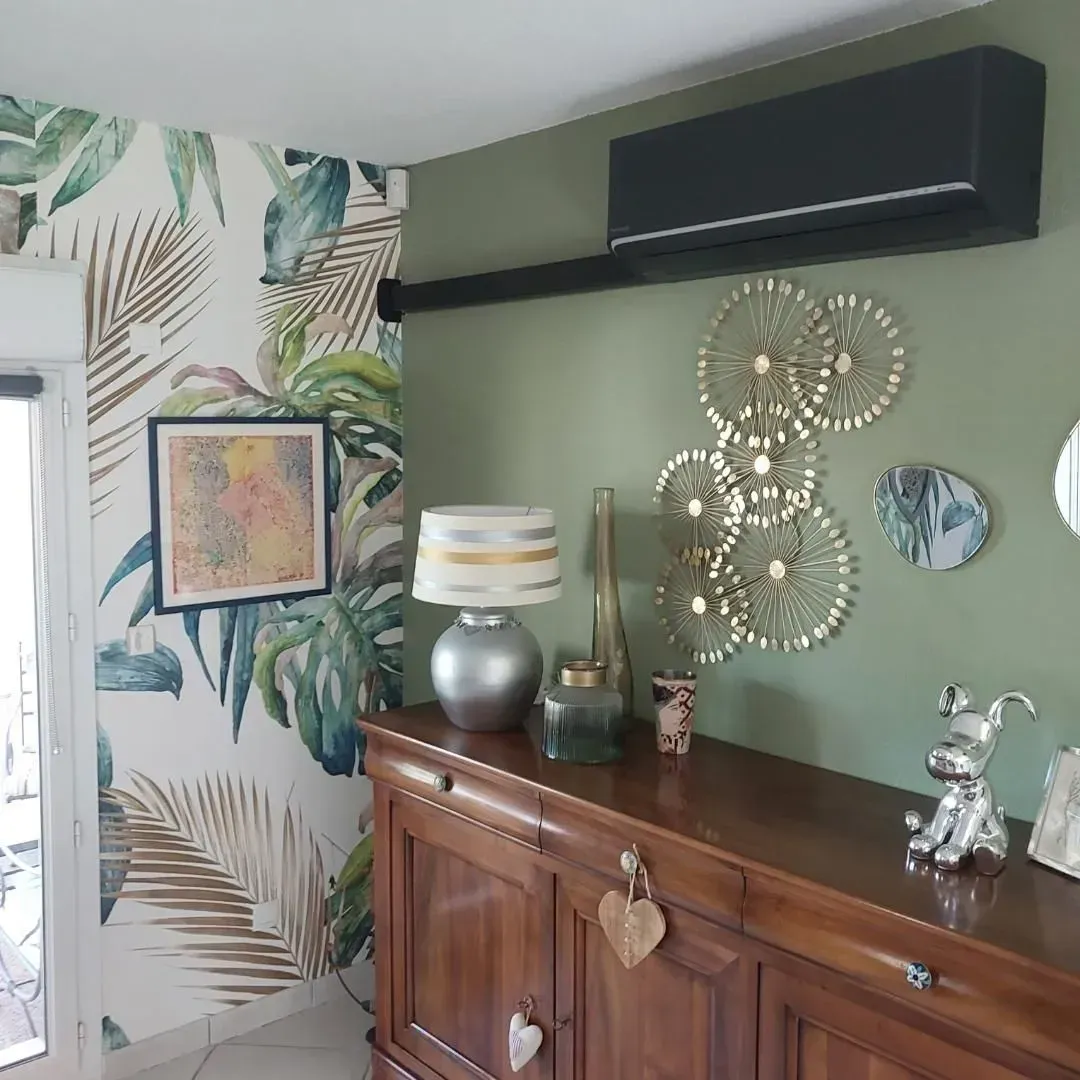 Un meuble en bois est adossé à un mur vert sauge, à côté d'un papier peint tropical orné d'éléments décoratifs, d'une lampe et d'un climatiseur noir.