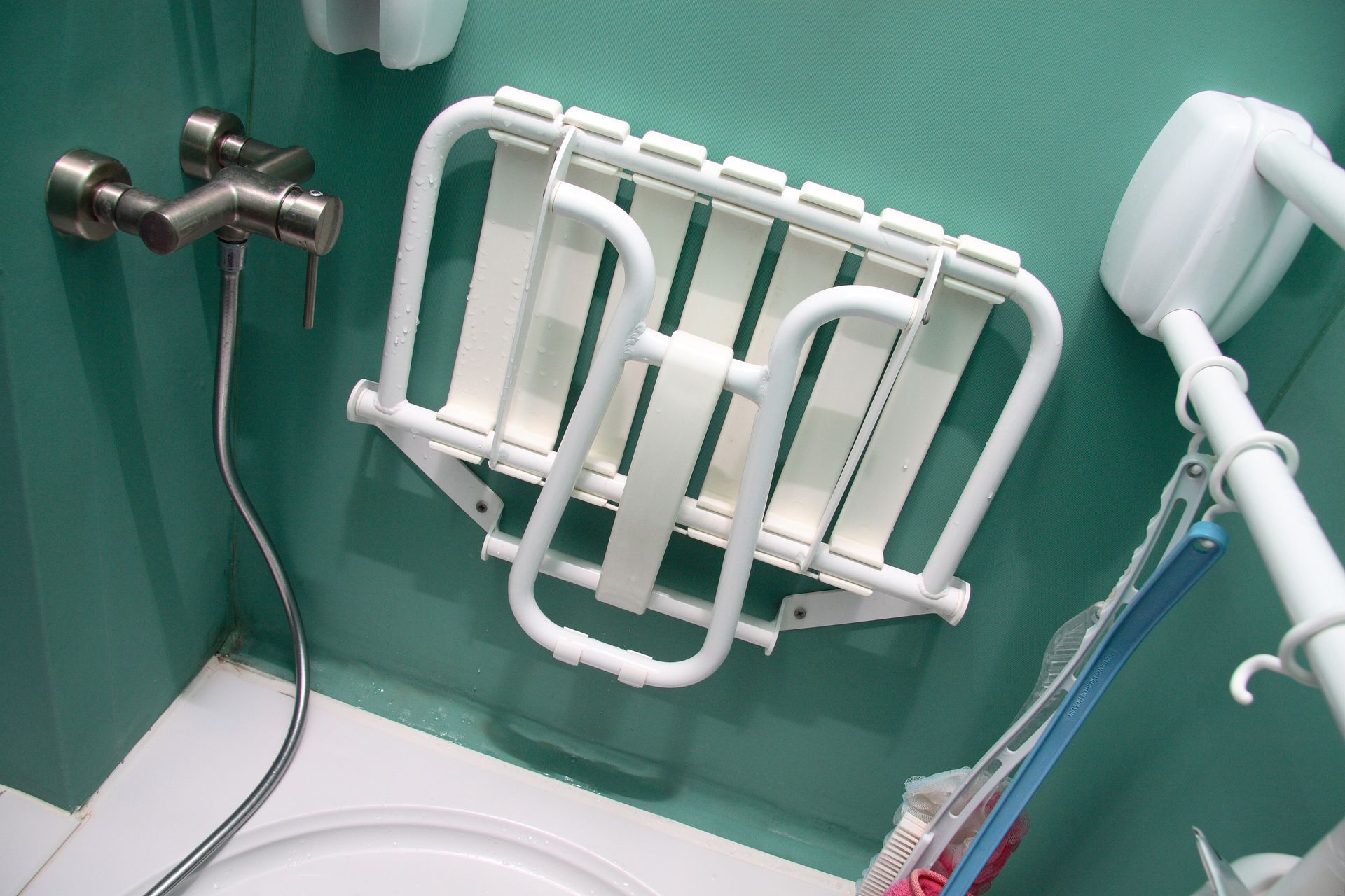 Siège de bain blanc installé dans une cabine de douche bleu sarcelle, avec des barres d’appui métalliques et une douchette à main.