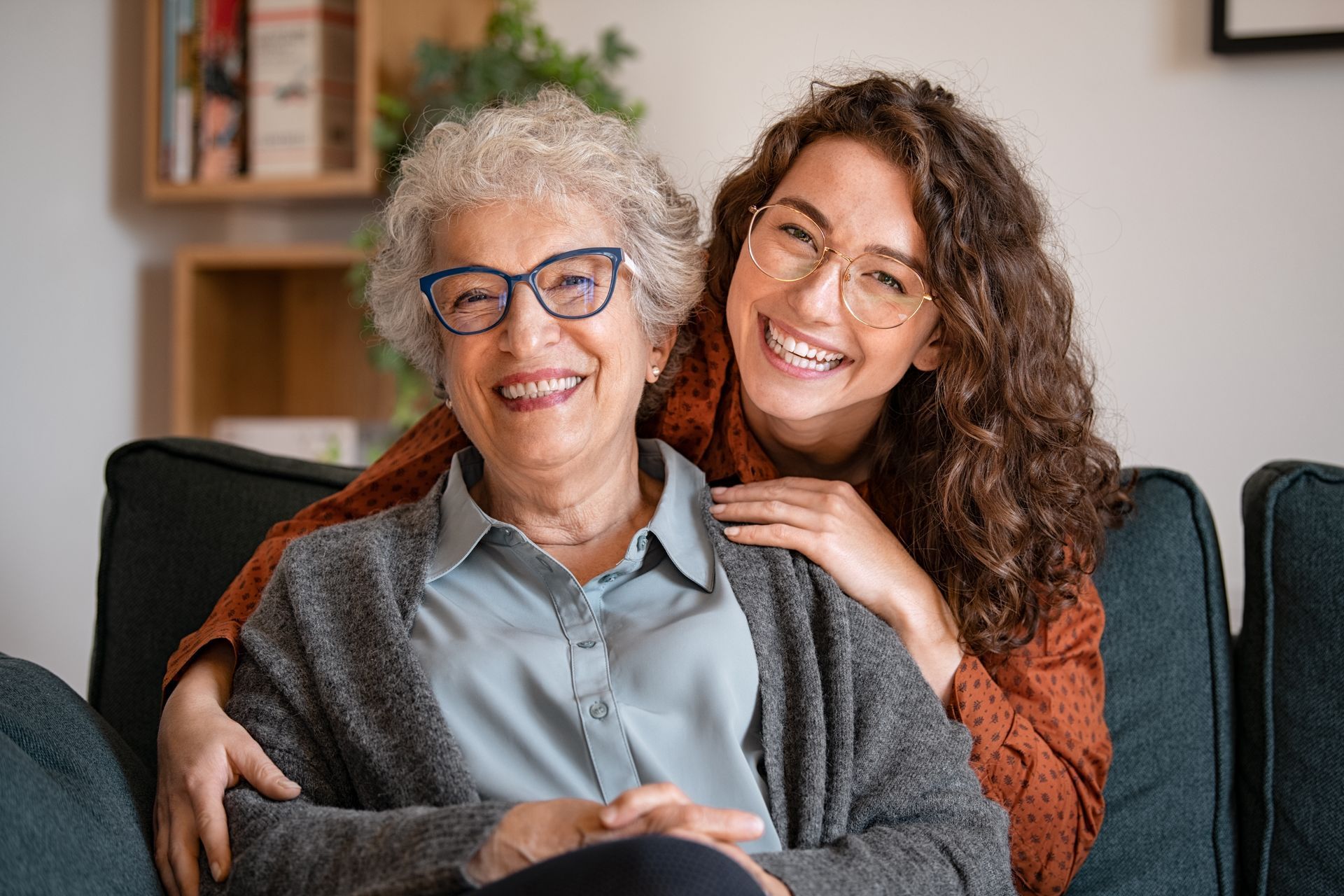 Deux femmes sourient sur un canapé, l’une ayant passé un bras autour de l’autre dans un salon chaleureux.