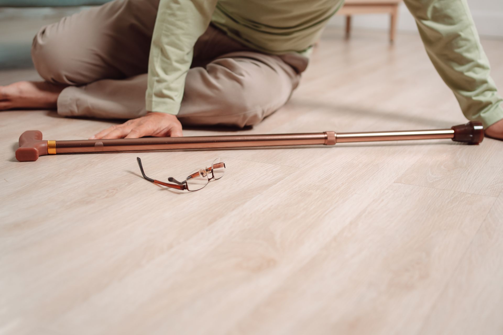 Personne agenouillée à côté d’une longue tige en bois posée sur un parquet clair, avec une paire de lunettes à proximité