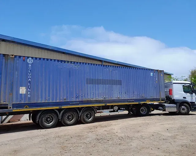 Un contenedor de envío azul está estacionado al lado de un camión blanco.