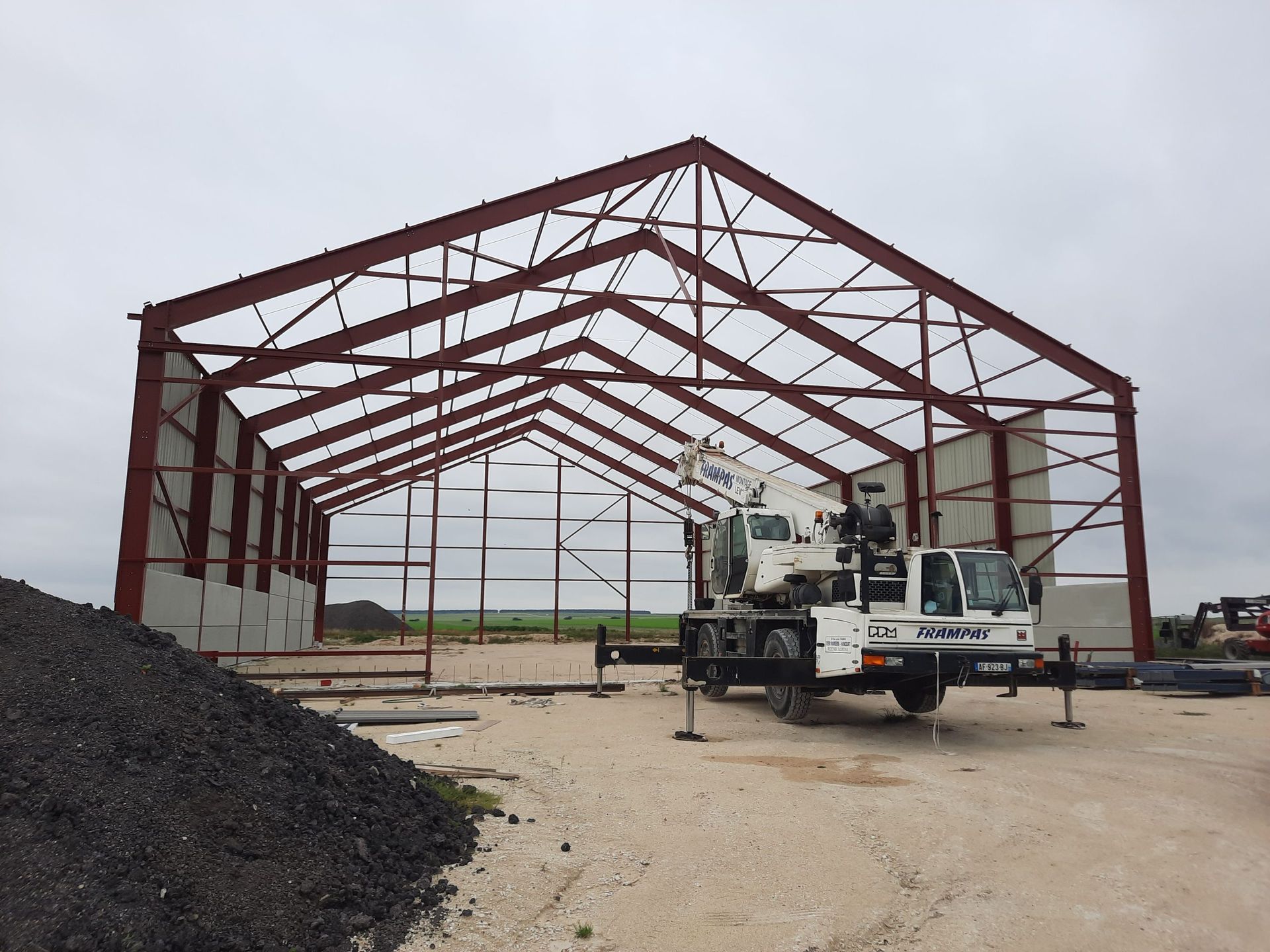 Bâtiment à ossature métallique en construction, avec une grue sur un terrain de gravier sous un ciel nuageux.