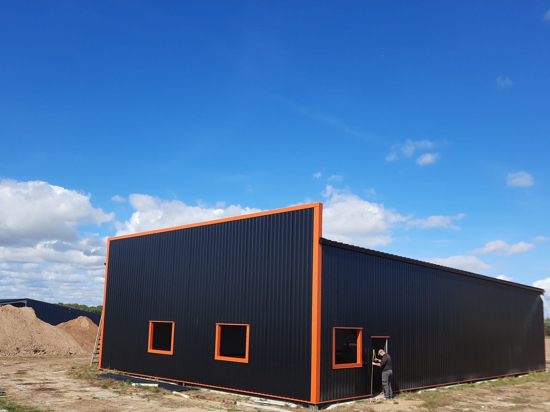 Bâtiment noir aux boiseries orange et à trois fenêtres se détachant sur un ciel bleu parsemé de nuages. Une personne se tient à proximité.