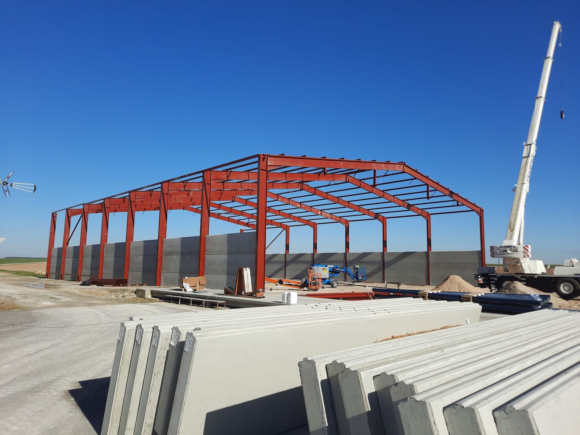 Bâtiment à ossature métallique en construction, avec une grue, sur fond de ciel bleu. On aperçoit des blocs et des panneaux de béton.