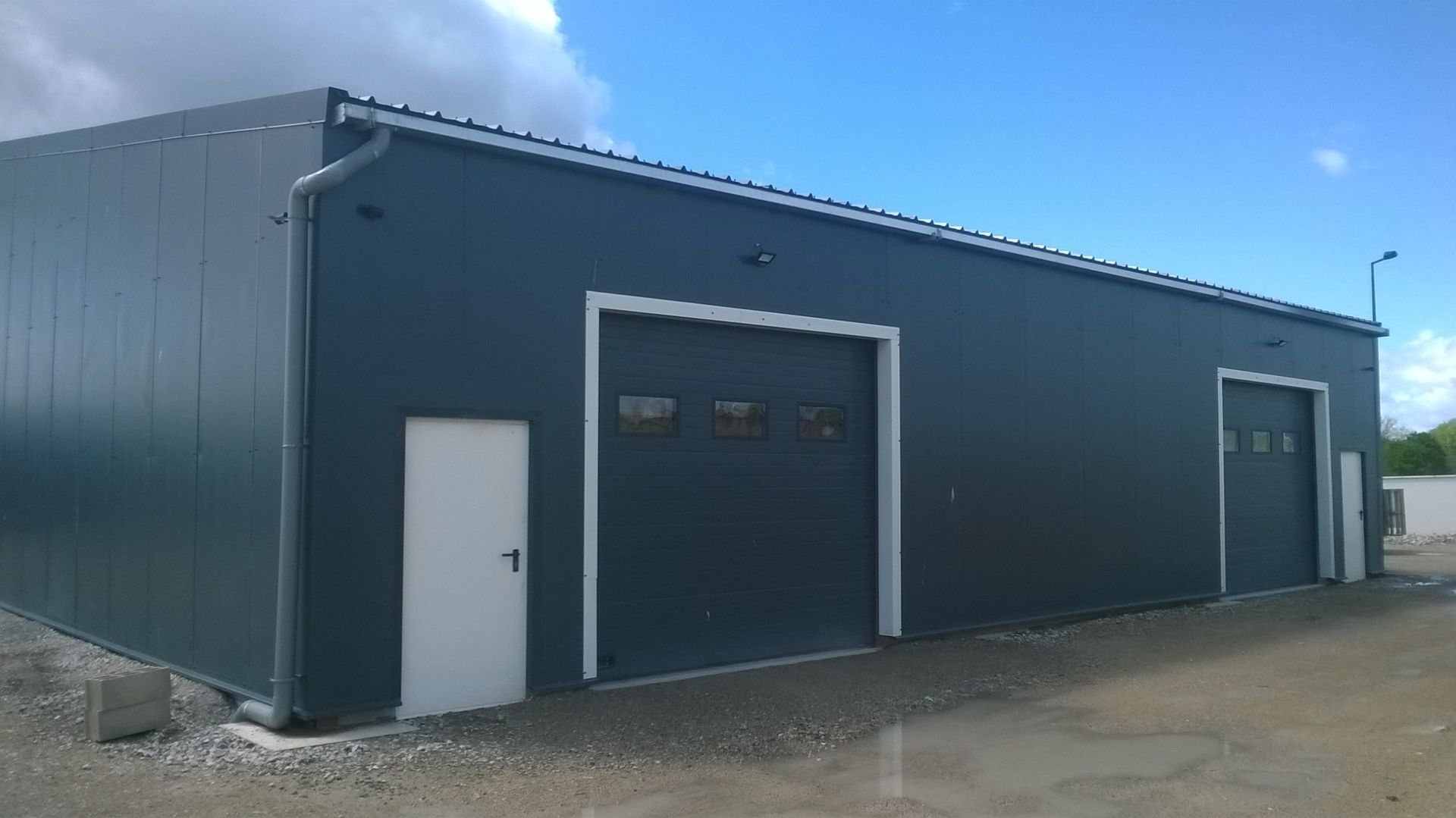 Bâtiment industriel gris avec portes de garage et porte blanche, sous un ciel bleu.