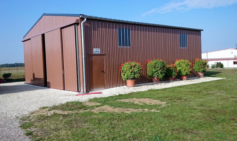 Abri de jardin en métal marron avec sol en gravier, fleurs rouges en pot et ciel bleu.