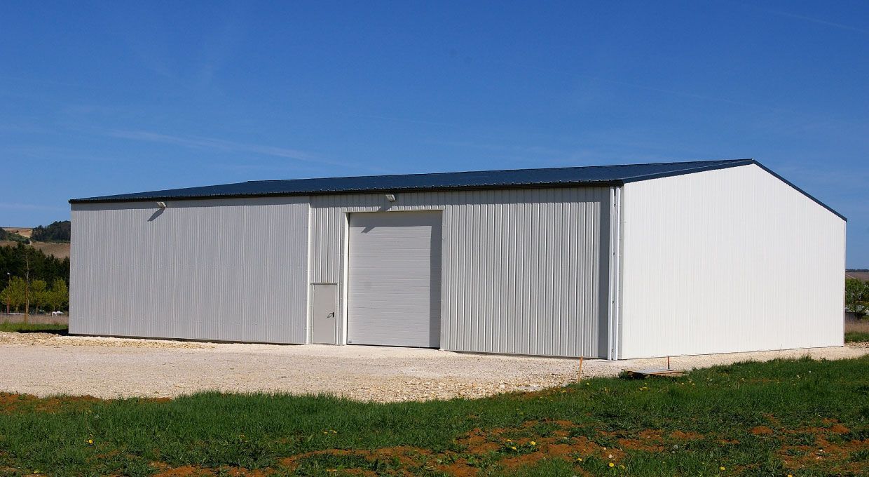 Bâtiment de stockage en métal blanc avec un toit sombre, sur un sol en gravier, sous un ciel bleu.