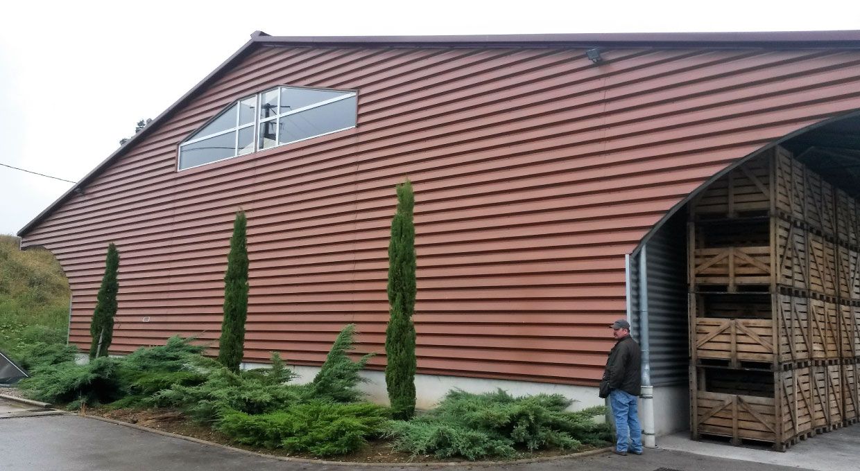 Bâtiment brun avec une fenêtre cintrée, un homme près d'un entrepôt rempli de caisses en bois et des arbustes verts.