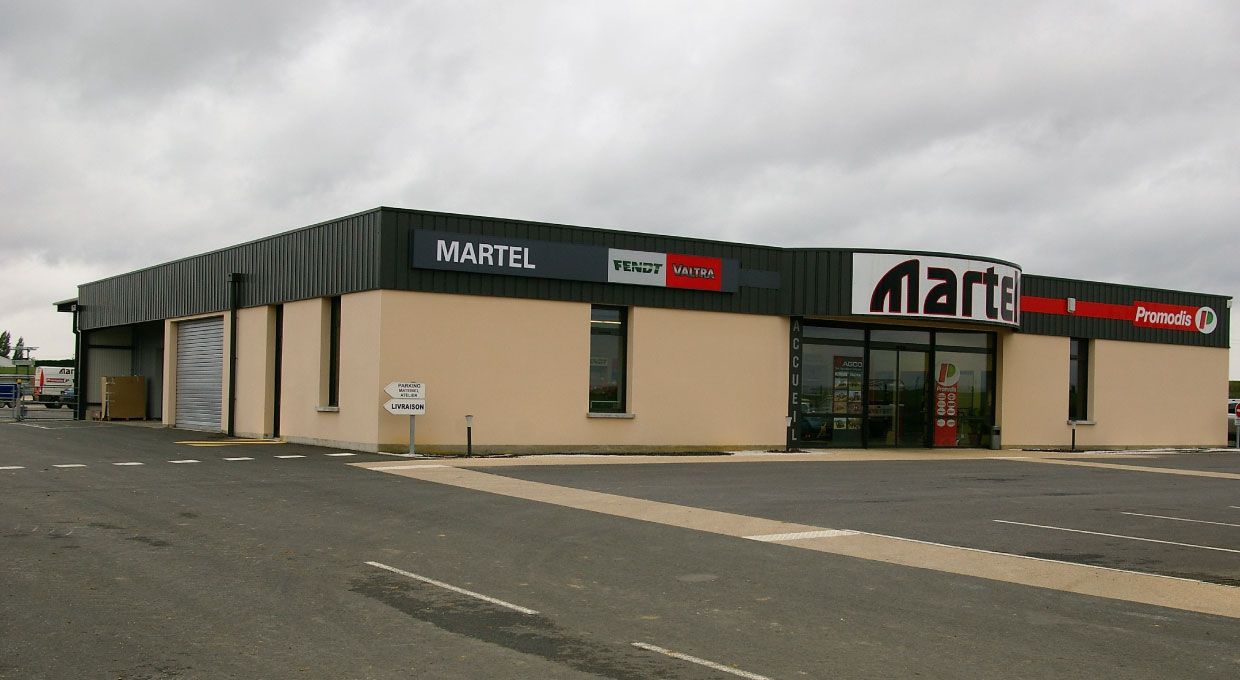 Un bâtiment beige et noir avec une grande enseigne Marti, sous un ciel couvert, avec un parking vide.