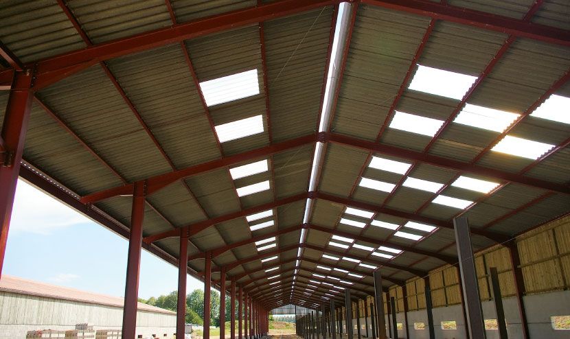 Vue intérieure d'un bâtiment à ossature métallique rouge avec un toit en tôle ondulée, doté de puits de lumière.