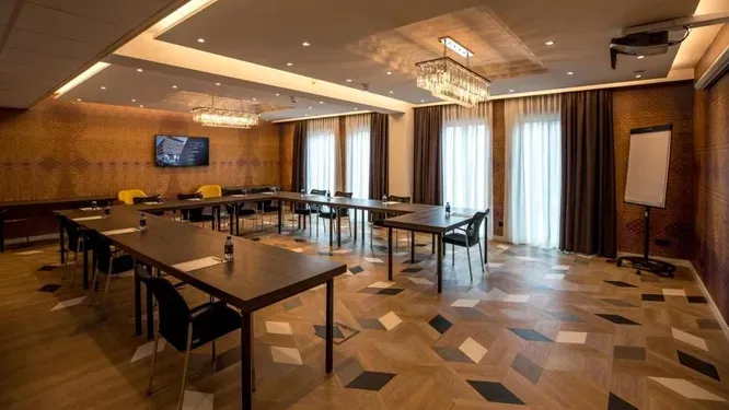 Een moderne, lege vergaderzaal in een hotel met U-vormige tafels, een houten vloer met patroon, kroonluchters en grote ramen.