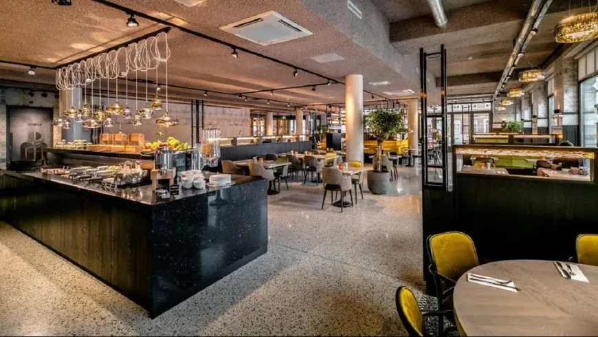 Een moderne, open ontbijtruimte in het hotel met een buffet van zwart marmer, terrazzovloeren en ronde tafels met gele stoelen.