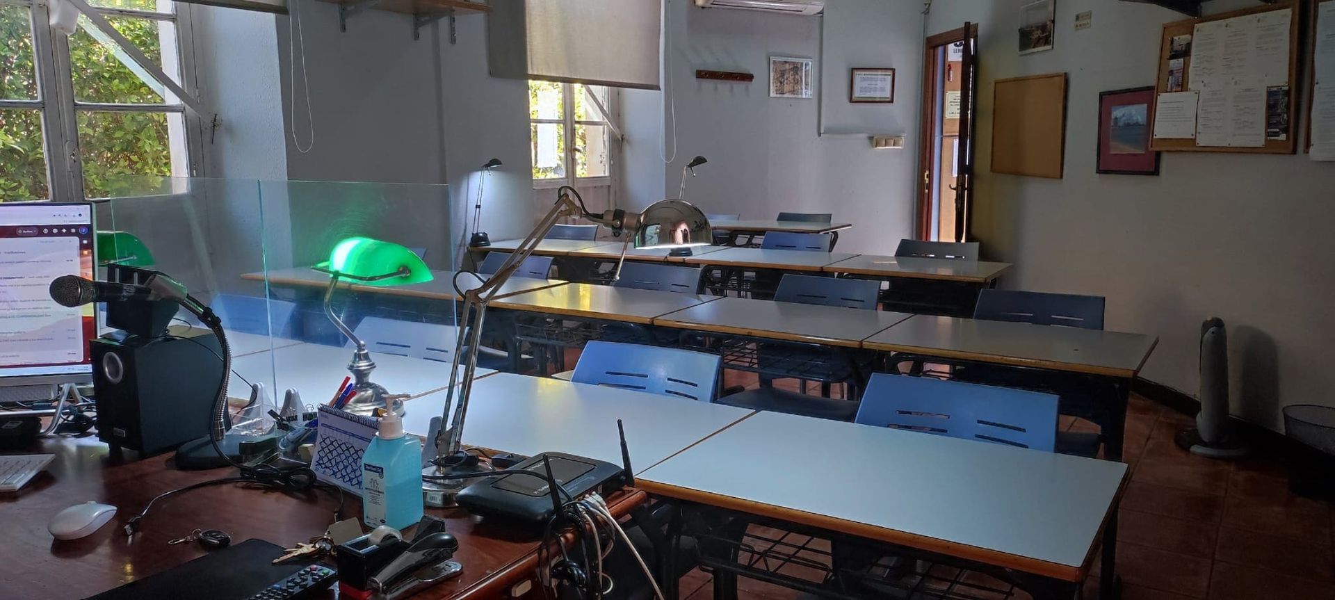 Un aula vacía con pupitres largos de color claro, sillas azules y un objeto de color verde brillante sobre una mesa.