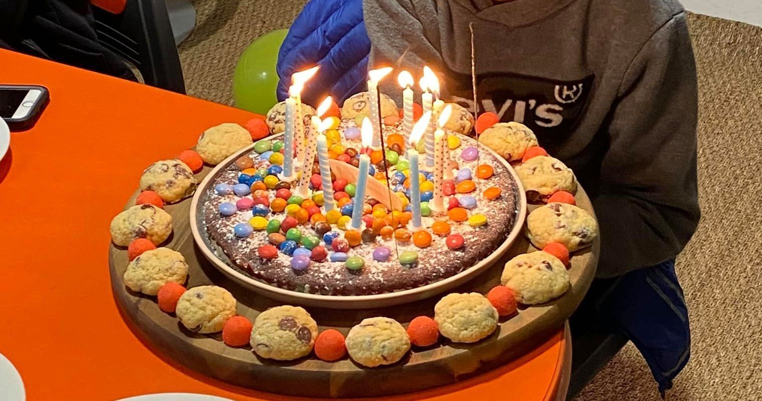 Gâteau d'anniversaire avec des bougies allumées, garni de bonbons et de biscuits, sur une table orange.