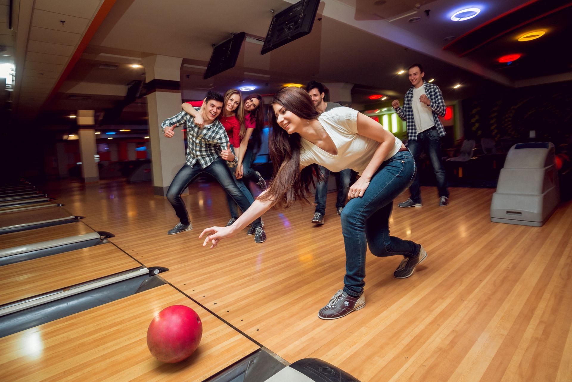 Une femme joue au bowling et lance une boule rouge sur la piste, tout sourire. Des amis applaudissent en arrière-plan, au bowling.