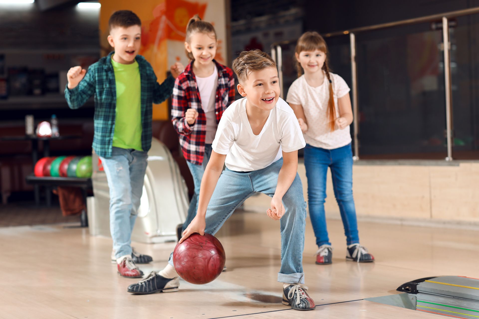 Quatre enfants jouent au bowling ; un garçon est sur le point de lancer la balle, les autres enfants applaudissent dans la salle de bowling.