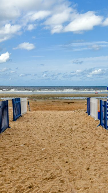 Chemin de sable menant à la plage et à la mer sous un ciel bleu avec des nuages blancs.