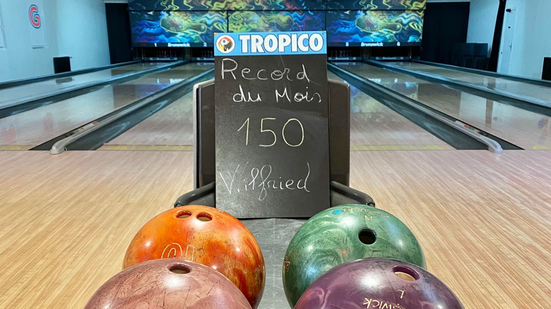 Boules de bowling au premier plan, pistes en arrière-plan. Panneau avec Record du Mois de noter et logo Tropico.